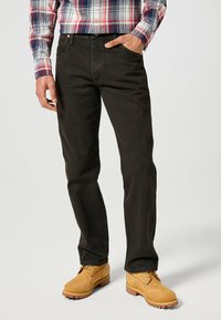 Pantalons en denim noir pour homme avec une coupe droite. Portés avec des bottes de travail beiges et une chemise à carreaux rouge, bleue et blanche. Possède des poches avant.
