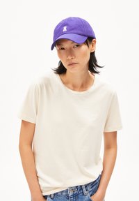 Lila Baumwollmütze mit einem kleinen gestickten Logo vorne, kombiniert mit einem hellbeigen T-Shirt und Jeans.