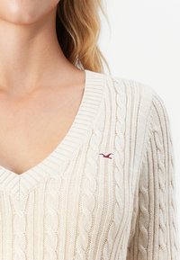 Sieviete, kas valkā krēmkrāsas trikotāžas V veida kakla džemperi ar nelielu sarkanu izšūtu putnu logotipu uz krūtīm.