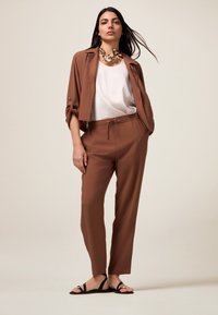 Veste courte marron portée sur un chemisier blanc, associée à un pantalon marron avec cordon de serrage et des sandales noires avec des accents dorés. Textures et couleurs simples.