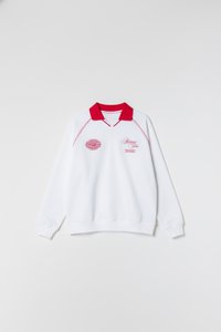 Λευκό φούτερ με κόκκινο γιακά, διακοσμημένο με ροζ κεντημένα λογότυπα και κείμενο στο εμπρός μέρος. Μακριά μανίκια με ριμπ μανσέτες και τελείωμα.