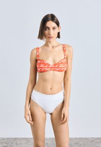 Czerwony top bikini z białymi napisami, z szerokimi ramiączkami i zakrzywionym kształtem; połączony z białymi majtkami z wysokim stanem. Gładka faktura.