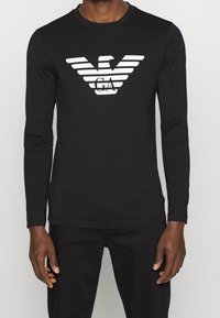 Svart långärmad t-shirt med en framträdande vit örnlogotyp och horisontella ränder över bröstet, tillverkad av ett slätt, elastiskt tyg.