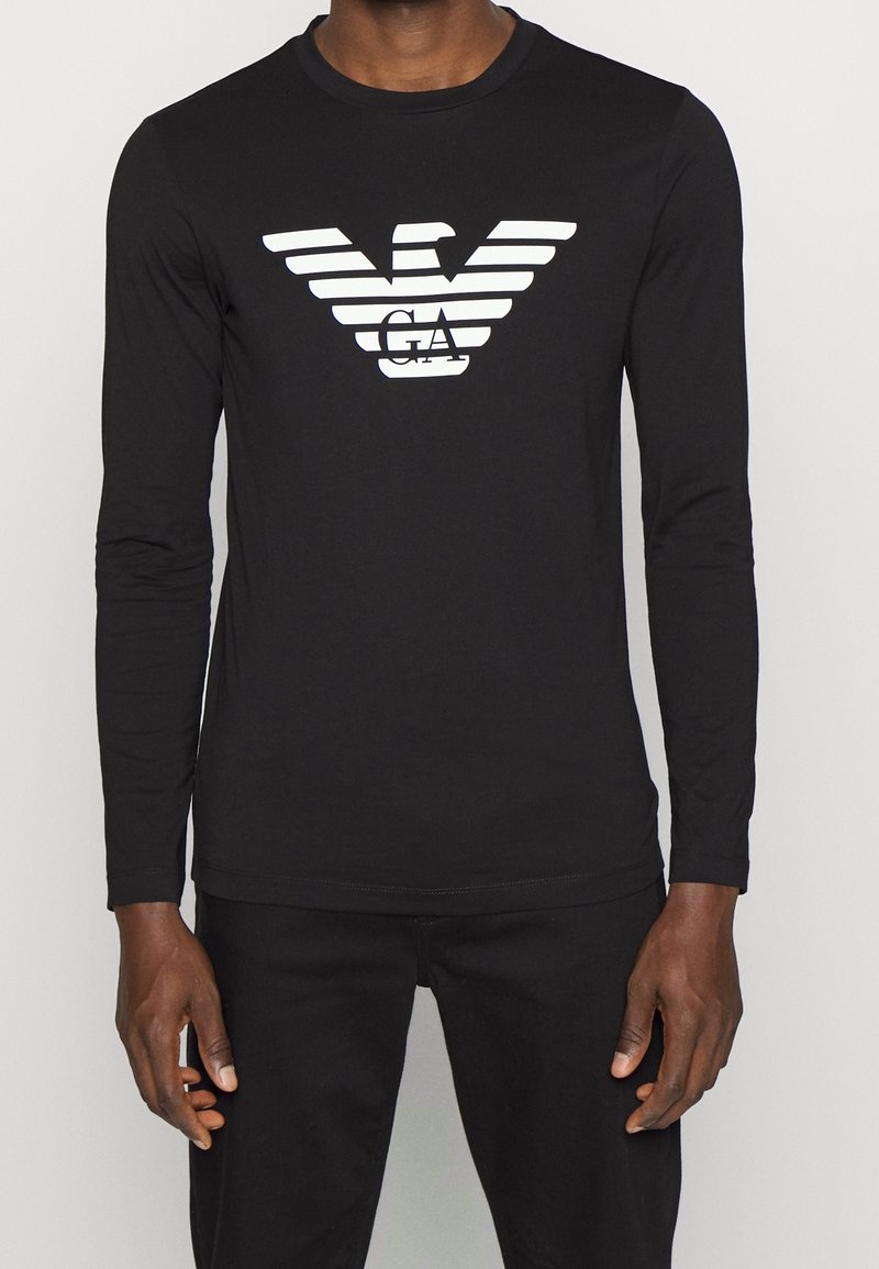 Svart långärmad t-shirt med en framträdande vit örnlogotyp och horisontella ränder över bröstet, tillverkad av ett slätt, elastiskt tyg.