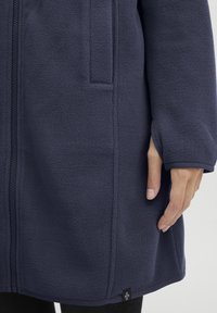 Chaqueta de forro polar azul marino con textura suave, cremallera frontal, bolsillos laterales y etiqueta de logo en el dobladillo. Mangas largas con puños ajustados.