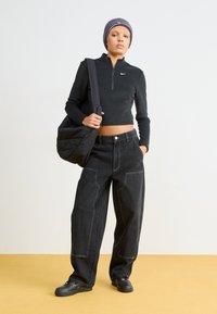 Čierny zipsový top s dlhým rukávom, voľné čierne džínsy s viditeľným švom a čierne topánky. Modelka nesie textúrovanú čiernu tašku.