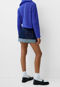 Pull violet coupé et texturé, associé à une jupe en denim foncé avec un ourlet bleu clair et des coutures rouges, porté avec des chaussettes blanches et des mocassins.