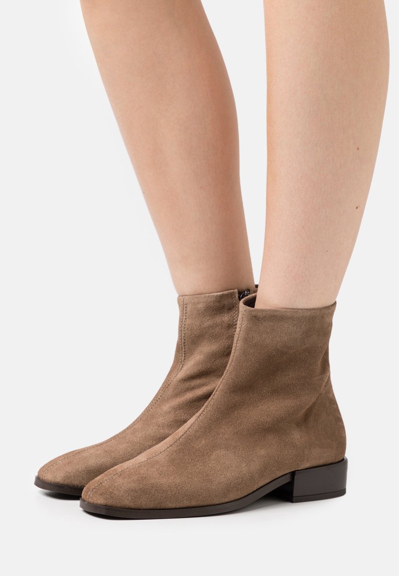 Bottines en daim marron avec un talon bloc, présentant un design épuré et élancé, ainsi qu'une fermeture éclair sur le côté intérieur. Texture lisse.