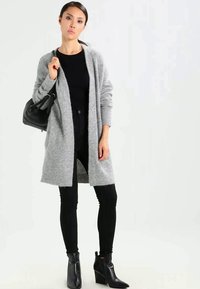 Cardigã longo de frente aberta em cinza claro, com textura de malha suave. Usado sobre uma blusa preta e calças skinny pretas, acompanhado de uma bolsa preta.