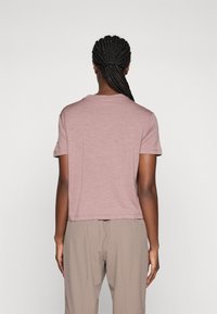 Person med flette i håret som er samlet bakover, iført en lys mauve kortermet skjorte og beige bukser, snur ryggen mot en ensfarget bakgrunn.