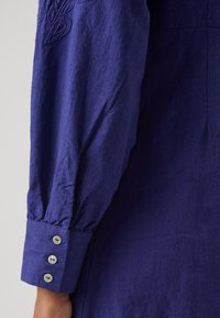 Manica di camicia in lino viola con polsino arricciato e tre bottoni bianchi. Presenta dettagli ricamati nella zona della spalla.