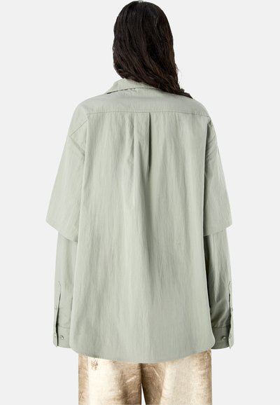 Persona con cabello largo y oscuro que lleva una camisa holgada verde salvia clara y pantalones beige, vista de espaldas sobre un fondo liso.