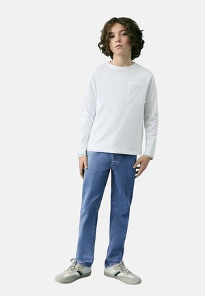 T-shirt blanc à manches longues avec une poche, associé à un jean bleu clair et des baskets grises avec des rayures vertes. Design simple et coupe décontractée.