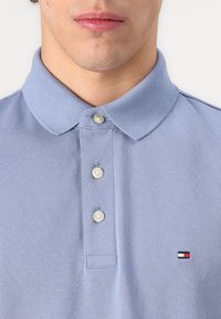 Mand iført en lyseblå polo med tre knapper og et lille rektangulært logo i rød, hvid og sort på brystet.