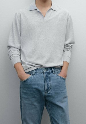 Personne portant un polo gris clair à manches longues rentré dans un jean bleu taille haute, mains dans les poches, sur un fond uni.