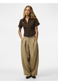 Femme aux longs cheveux blonds porte une chemise marron à manches courtes boutonnée et un pantalon large beige plissé, les mains dans les poches.