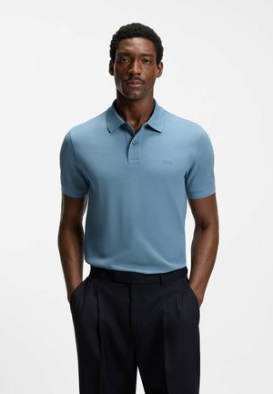 Man met kort krullend haar en snor, gekleed in een lichtblauw poloshirt met korte mouwen en een donkere broek, met handen in de zakken.