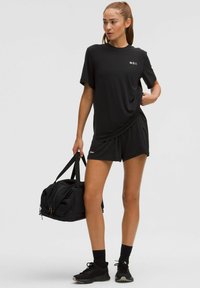 Fekete sportnadrágot, pólót és edzőcipőt viselő nő egy fekete sporttáskával áll egy egyszínű világos háttér előtt.