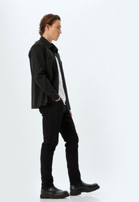 Camisa negra de botones sobre una camiseta blanca, combinada con jeans ajustados negros y botas negras de suela gruesa, mostrada desde un perfil lateral contra un fondo sencillo.