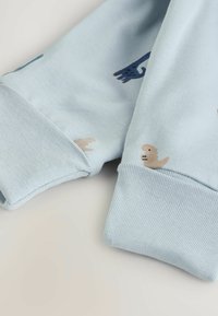 Helles blaues Sweatshirt mit gerippten Bündchen, das verspielte Tiermuster zeigt, darunter eine beige Robbe und dunkelblaue Designs auf dem Stoff.