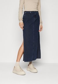 Mörkblå denim maxikjol med en sidoklyv, i kombination med beige sneakers. Kjolen har klassisk sömnad och en traditionell femfickdesign.