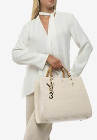 Sac beige avec poignées en bambou, motif embossé, quincaillerie dorée et un porte-clés décoratif avec le numéro 3.