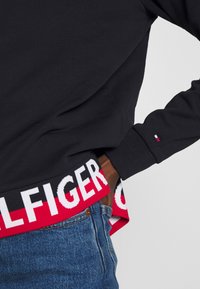 Felpa nera con un elastico a marchio rosso e bianco, caratterizzata da un logo e un tessuto testurizzato, abbinata a jeans in denim blu.