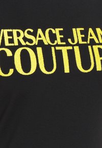 Tissu noir avec un texte jaune brodé en gras indiquant Versace Jeans Couture.