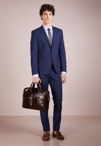 Abito blu marina con un design aderente, abbinato a una camicia a righe azzurro chiaro e una cravatta grigia. Tiene in mano una valigetta in pelle marrone con dettagli blu.