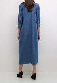 Denim-mekko, jossa on kolme neljäsosaa hihat, sivuhalkiot ja suora siluetti. Väri: keskisininen. Yhdistetty mustiin korkokenkiin.