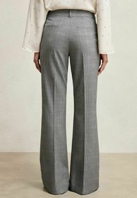 Femeie îmbrăcată cu pantaloni largi în carouri gri și o bluză albă cu buline negre, stând cu spatele pe un fundal neutru.