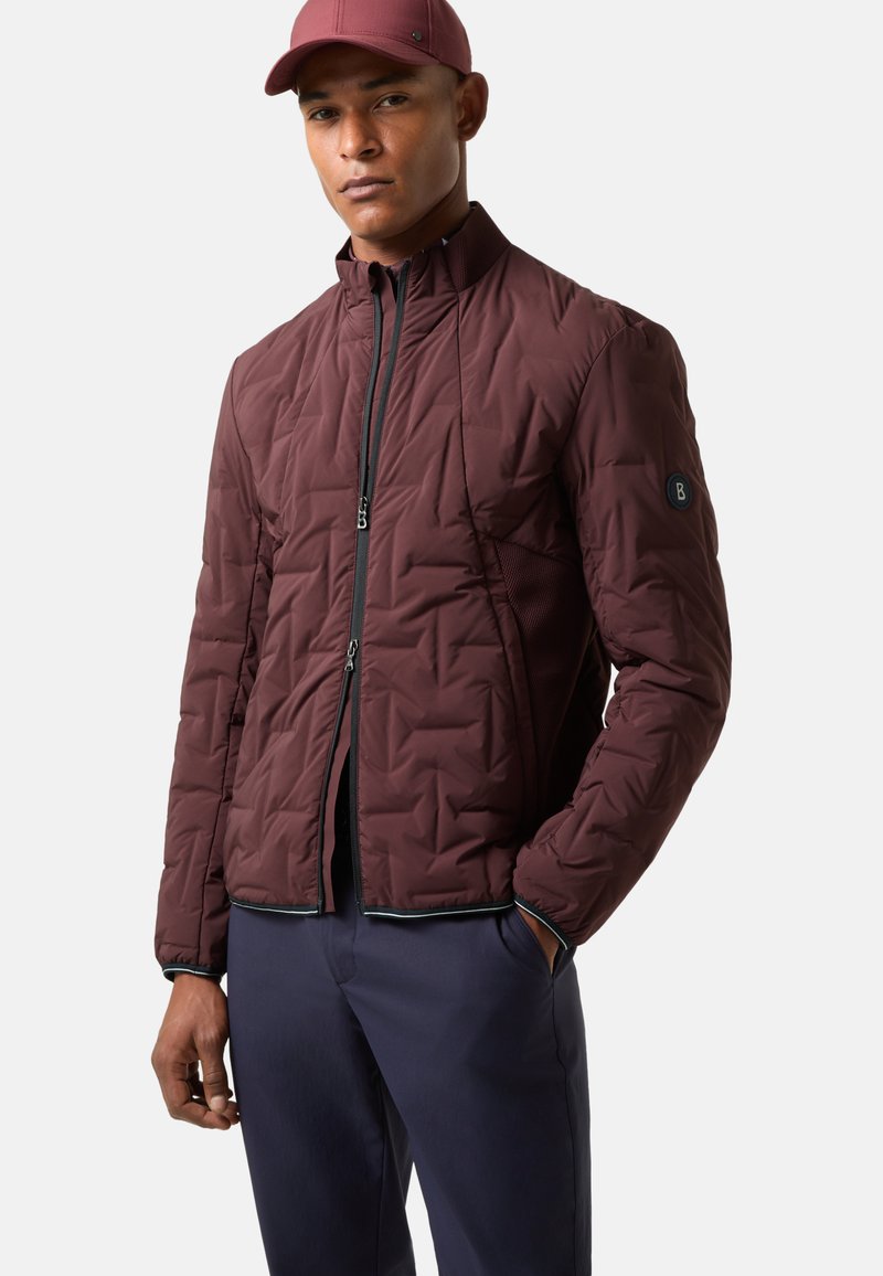 Bordeauxroter Stepp-Puffer mit gesteppter Verarbeitung und Reißverschluss vorne. Mit kontrastierendem Besatz und einem Logo-Patch am Ärmel. Wird mit dunkler Hose getragen.