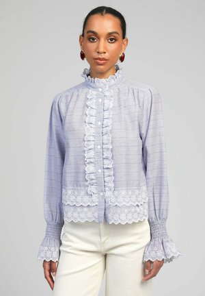 Femme portant un chemisier bleu clair à rayures avec des volants en dentelle blanche sur le col, le devant, les poignets et l'ourlet, associé à un pantalon écru.