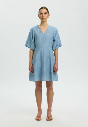 Kobieta stojąca przodem, ubrana w fakturowaną jasnoniebieską sukienkę z dekoltem w serek, bufkami na rękawach i beżowe sandały na jednolitym tle.