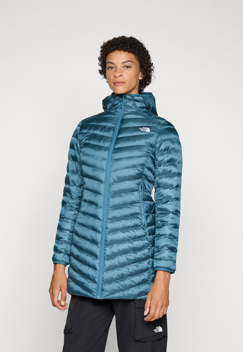 Teal isolert jakke med horisontalt quiltet mønster, høy krage, hel glidelås og logo på brystet. Glatt stoff og lange ermer.