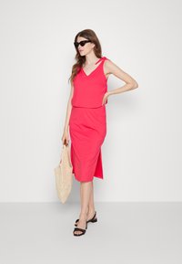 Vestido rosa sem mangas com decote em V, fenda lateral e corte descontraído. Combinado com sandálias pretas e uma bolsa de palha.