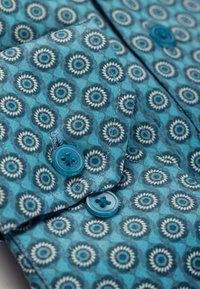 Nahaufnahme eines blau gemusterten Stoffs mit kreisförmigen Motiven und zwei blau genähten Knöpfen nahe dem Rand.