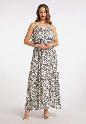Donna che indossa un vestito lungo bianco senza maniche con stampa leopardata nera e tacchi bianchi aperti davanti, in piedi davanti a uno sfondo bianco semplice.