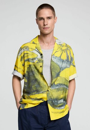 Mann trägt gelbes, grau abstrakt gemustertes kurzärmeliges Hemd über grauem Tanktop, mit marineblauer Hose, Hände in den Taschen, neutraler Hintergrund.