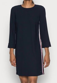 Navyblaues Kleid mit Dreiviertelärmeln, versehen mit seitlichen Streifen in Rot, Weiß und Blau. Glattes Material und knielanges Design.