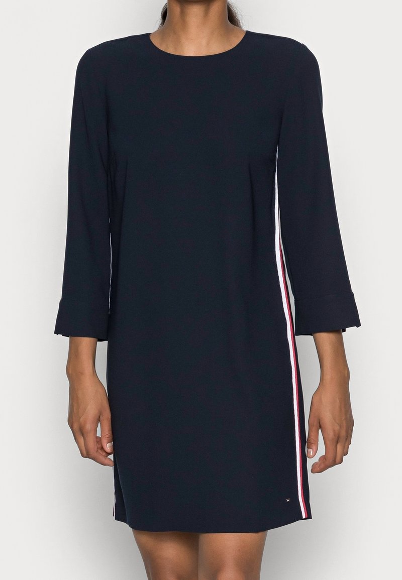 Navyblaues Kleid mit Dreiviertelärmeln, versehen mit seitlichen Streifen in Rot, Weiß und Blau. Glattes Material und knielanges Design.