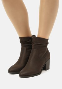 Botas tobilleras marrones con un diseño arrugado, material de imitación de cuero suave, punta afilada y tacón cuadrado cónico. Detalle de cremallera en la parte posterior.