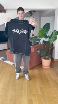 T-shirt preta com texto gráfico branco, mangas curtas e costura em contraste. Segurada por uma pessoa num espaço habitacional com plantas.