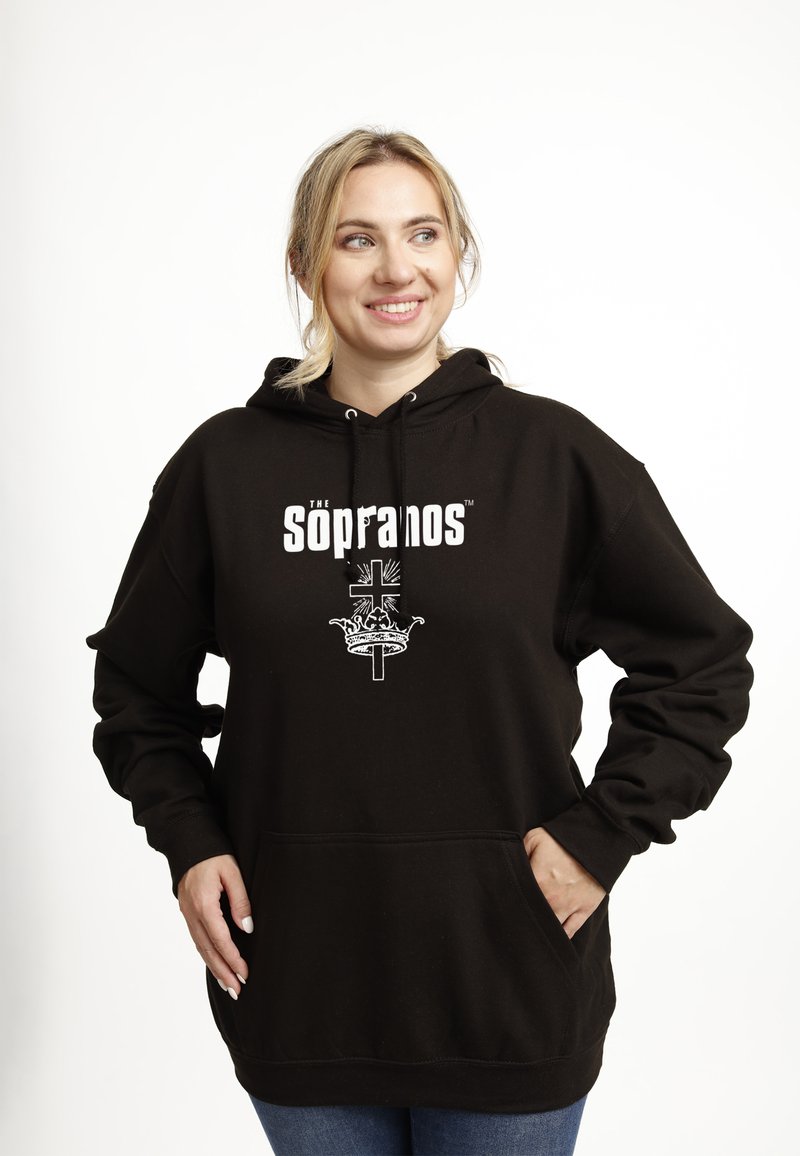 Donna sorridente con capelli biondi in una felpa nera "I Soprano" con un design di croce bianca e corona, mani nella tasca anteriore, che guarda a destra.
