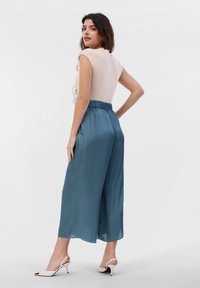 Light beige fitted top with cap sleeves; teal satin wide-leg pants with elastic waistband; model wearing white heeled mules.