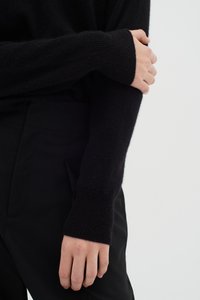 Pulover negru tricotat cu mâneci lungi, prevăzut cu manșete rib, textură moale, asortat cu pantaloni negri croiți, parțial vizibili.