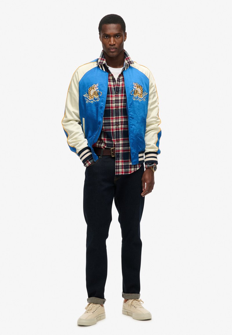 Veste bomber en satin bleu avec des manches blanches, design de tigre brodé, chemise à carreaux, jean en denim foncé et baskets beiges.