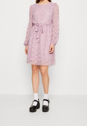 Frau trägt ein knielanges, rosa Spitzenkleid mit langen Ärmeln und einem gebundenen Taillengürtel, kombiniert mit weißen Socken und schwarzen Plateau-Schuhen.