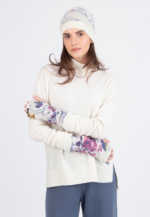 Beige Strickpullover mit hohem Kragen, kombiniert mit mehrfarbigen, blumigen Armschützern und einer passenden Mütze im gleichen Muster. Strukturierte, weiche Materialien.