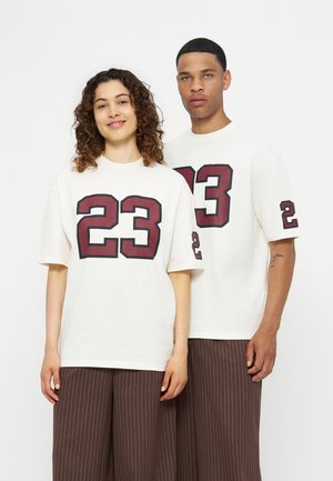 Deux mannequins sont côte à côte portant des chemises blanches oversize avec les grands numéros bordeaux 23 et 2 sur les manches, associés à un pantalon à rayures marron.